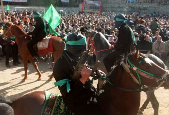 A masked Hamas gunman on horseback with other masked Hamas fighters on hoseback and a large croud nearby