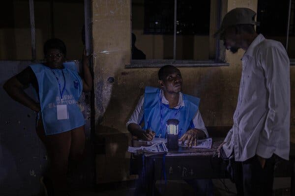 A person speaks to a polling station worker, who is writing while seated at a table. Another worker stands nearby.