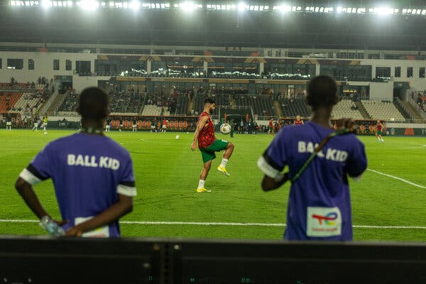 A soccer player bounces a ball on his knee in a stadium as two people wearing purple shirts that read “ball kid” on the back look on. 