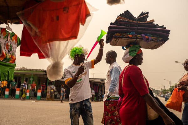 A market vendor wearing a green-and-white curly wig blows into a long, plastic horn. Soccer clothing hangs from his stall while people walk by.