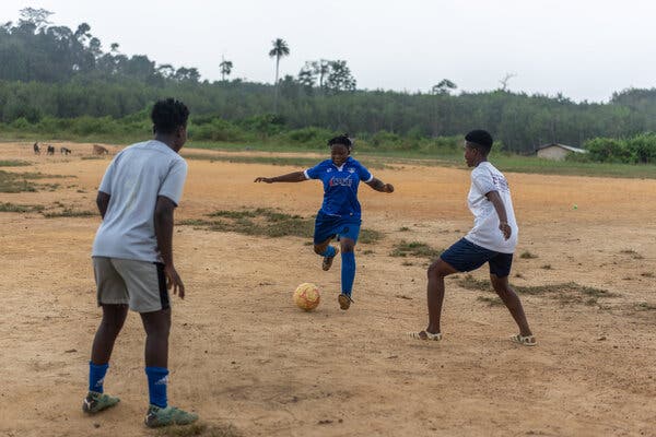 Three soccer players kick around a ball on a dirt field.