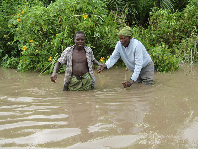 Two men stand in floodwaters above their knees, the younger man holding the arm of the older man, who is leaning on a cane in the water.