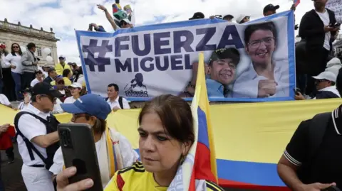 EPA People demonstrate with signs and flags during the March of Silence in Bogota, Colombia, 15 June 2025. A woman, wearing the shirt of Colombia's national football team, can be seen holding up a mobile phone. Behind her, people are carrying a banner reading Strength, Miguel. 